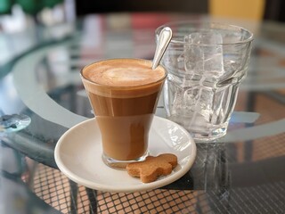 Coffee with milk, a cookie and a glass with ice on a glass table.