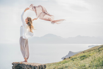 Young woman with long hair stands on coastline rocky cliff edge and holds shawl waved by strong...