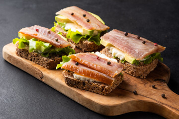 Sandwich with smoked fish, avocado, cream cheese, leaves salad  on rye bread. Snack on a dark wooden table.