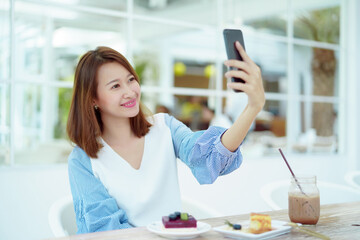 Portrait A beautiful Asian woman in a white shirt sits happily using her cell phone to take a selfie in a bakery.