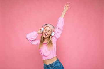 Photo of attractive positive smiling young blonde woman wearing pink blouse and pink hat isolated over pink background wall wearing white wireless bluetooth earphones listening to music having fun