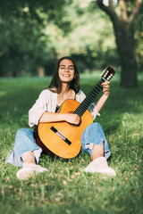 A laughing girl plays the guitar and sits on the grass in the park. Hobbies, music, musician