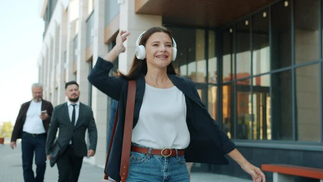 Happy young business lady is dancing walking outdoors wearing headphones outdoors in busy street. Leisure time and carefree businesspeople concept.