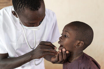 Cute little African boy opening his mouth compliantly while being tested for corona virus by a...