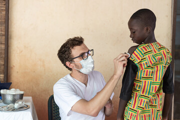 Young white volunteer preparing a small African boy to get a vaccine shot during childhood disease...