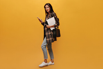 Attractive brunette woman in jeans and oversized jacket looks into camera, moves, holds phone and notebooks on yellow isolated background.