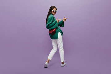 Fototapeta na wymiar Stylish woman in sunglasses, green sweaterwhite pants and sneakers with claret handbag dances on purple background. Happy girl moves, also points at some place