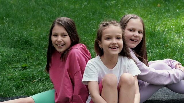 three beautiful happy tween teenagers girls in colorful clothes sitting on green grass in park. summertime. happy children. holidays, summer break. sisters, best friends, bff, sisterhood. slow motion.