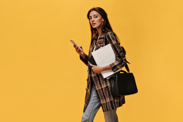 Young brunette woman in oversized jacket and jeans holds phone and white notebooks. Attractive girl with black bag poses on orange background.