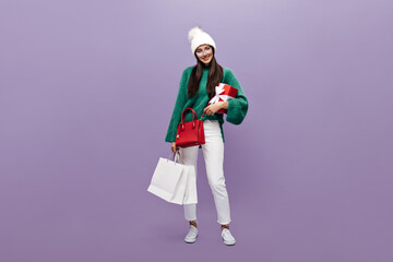 Long-haired brunette woman in white pants, green woolen sweater and warm hat smiles, holds red gift box and shopping bags on purple background.