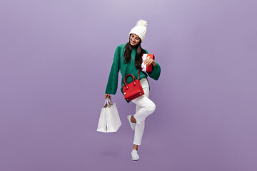 Brunette woman in white pants and green sweater moves on purple background. Charming girl in warm hat holds red gift box and shopping bags on isolated.