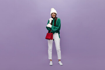 Brunette girl in stylish woolen sweater and white pants smiles on isolated. Cool long-haired woman on green top, knitted hat and gloves poses with red handbag on purple background.