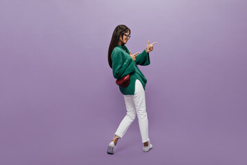 Fototapeta na wymiar Pretty woman in stylish glasses, green woolen sweater and white pants points at place for text on purple background. Cheerful girl stands with her back but turned to the right