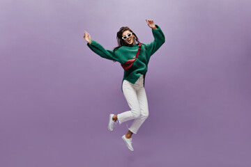 Fototapeta na wymiar Cheerful woman in white pants and stylish oversized green sweater moves on purple background. Happy brunette girl in sunglasses jumps on isolated.