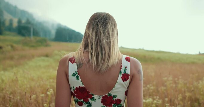 Girl in the white dress with red flowers, filmed from the back walking trough the field, touching ears of grass moving in the wind. Shallow depth of field, 4K