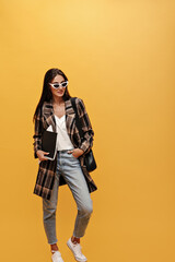 Young charming girl in denim pants, long oversized jacket and sunglasses poses on isolated. Brunette woman in stylish outfit holds notebooks on yellow background.
