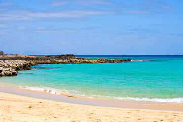 Obraz premium View to the horizon from the shore with white sand and rocks and transparent, sparkling blue ocean water in different shades. Fuerteventura, Canary Islands.
