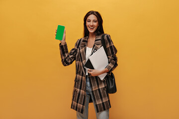 Cheerful girl in coat and jeans smiles and holds notebooks. Pretty brunette woman demonstrates screen of phone on yellow background.