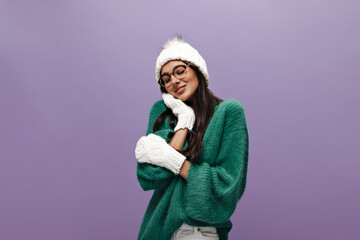 Charming woman in green sweater and eyeglasses poses on purple background. Young brunette girl in gloves and knitted hat smiles on isolated.