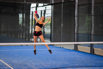 Woman playing padel in a blue grass padel court indoor - Young sporty woman padel player hitting ball with a racket