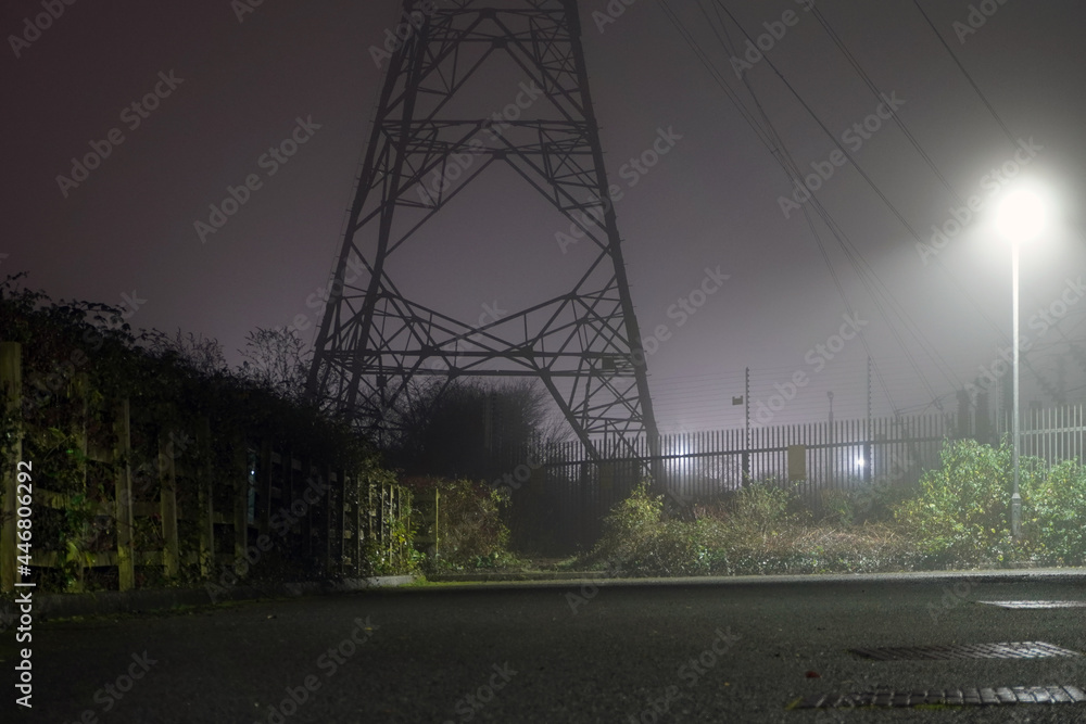 Poster an electricity pylon on an industrial estate back lighted on a foggy winters night.