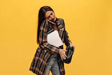 Brunette long-haired woman holds black handbag. Dissatisfied girl in jeans and oversized jacket talks on phone and poses with notebooks on isolated yellow background.