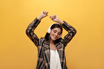 Joyful woman in white top, stylish brown jacket smiles and dances on yellow background. Attractive girl listening to music in headphones on isolated.