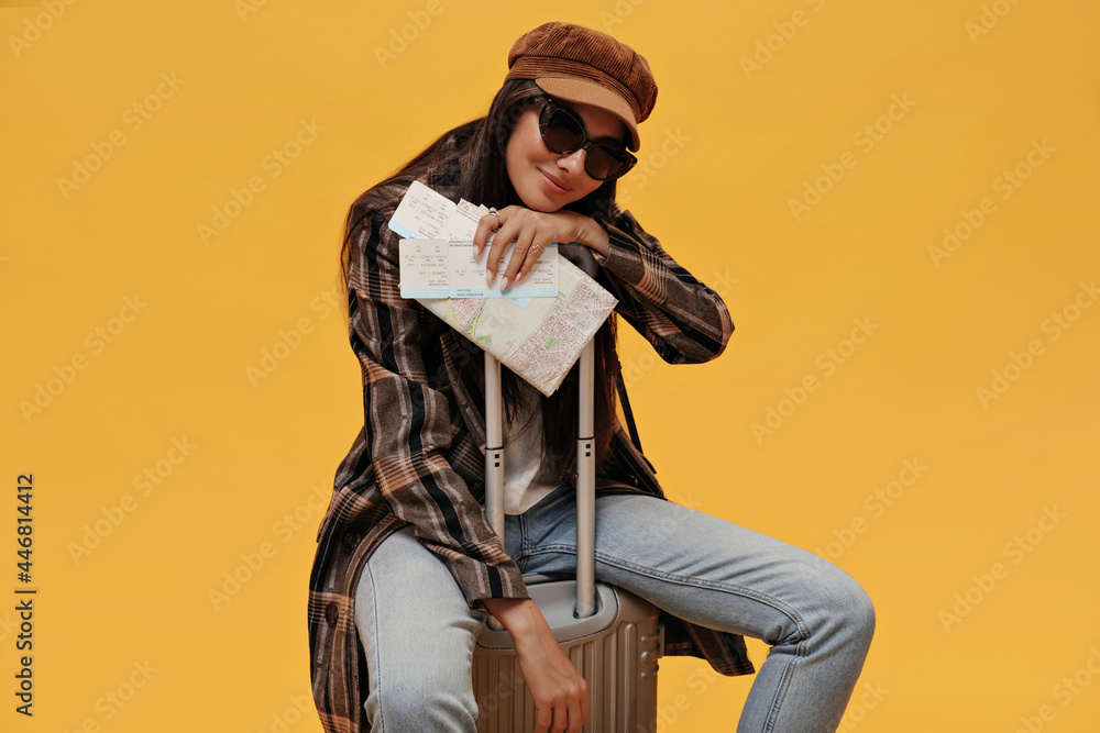 Wall mural Cool girl in sunglasses, beret and stylish coat smiles softly and sits on suitcase on isolated. Brunette woman in denim pants poses and holds map on yellow background.