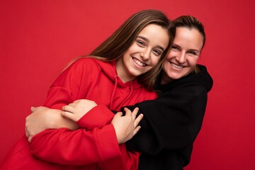 Photo shot of beautiful positive smiling brunette teenager wearing stylish red hoodie and adult brunette woman wearing trendy black hoodie standing isolated over red background wall looking at camera