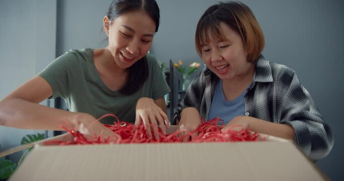 Happy young asia lady friends open cardboard delivery package from online marketplace in living room at home. Satisfied shopper surprise in internet unbox goods, Online shopping and delivery concept.
