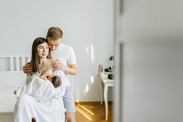 Obraz na płótnie Canvas Love couple sitting in cozy bedroom. Husband hug wife, woman breastfeeding a newborn baby. Mother holding daughter. Parents, motherhood. Family portrait concept. Trendy style.