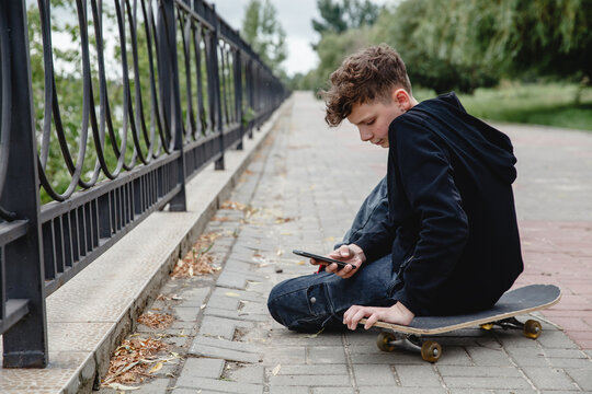 A curly-haired teen of European appearance in black hoodie sitting on an alley on a skateboard and talking on the phone