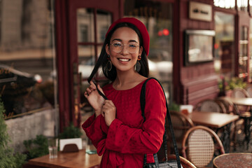 Charming brunette tanned woman in eyeglasses, red beret and stylish dress smiles widely and poses outside in street cafe.