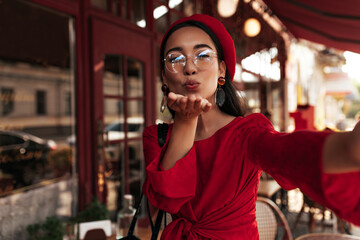 Happy Asian lady blows kiss outside. Cheerful brunette woman in beret and red dress takes selfie in street cafe.