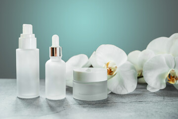 Fototapeta na wymiar White glass serum bottle and cream jar in the bathroom with orchid flowers in the background, unbranded cosmetic products, spa cosmetic product branding mockup