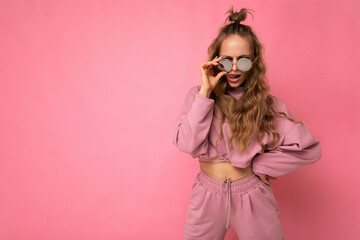 Attractive self-confident positive young blonde woman wearing everyday stylish clothes and modern sunglasses isolated on colorful background wall looking at camera