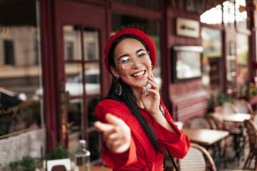 Good-humored Asian tanned woman in red beret, stylish dress and eyeglasses smiles and pulls hand to camera. Pretty brunette girl poses in street cafe.