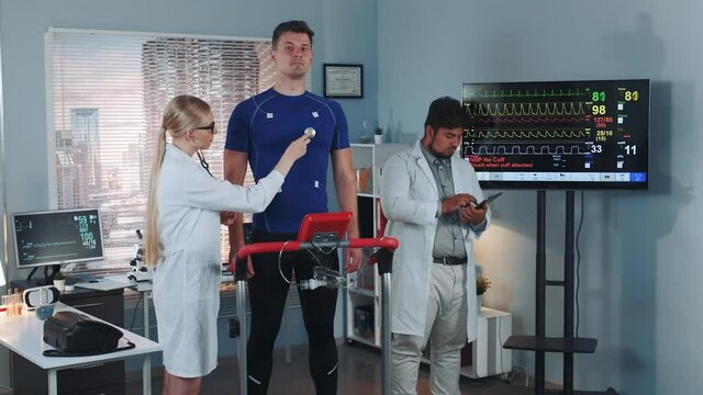 Female cardiology technologist listening to athlete heartbeat with a stethoscope while her colleague learning sportsman cardiogram. In Scientific Sports Laboratory.