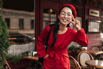 Cool tanned Asian woman in red dress and stylish beret shows peace sign. Attractive brunette lady in eyeglasses smiles in street cafe.