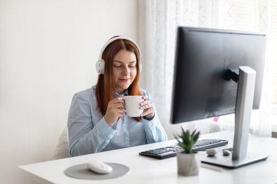 Young caucasian office woman worker sitting in wireless headphones with a cup of hot coffee or tea at the workplace at home or in the office - Powered by Adobe