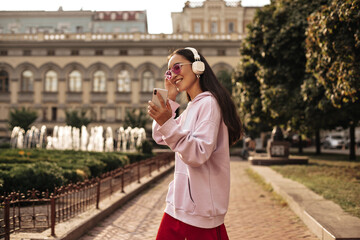 Pretty brunette Asian woman in pink sunglasses holds phone, smiles, listens to music in headphones. Happy girl in hoodie walks outside.