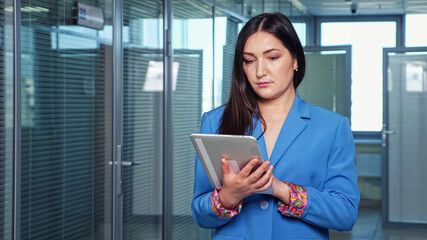 Long haired brunette businesswoman in blue jacket swipes grey tablet standing in premises lounge among offices with transparent glass doors close view