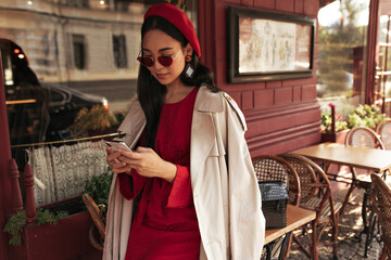 Pretty brunette woman in red dress, stylish sunglasses, beret and beige trench coat holds cellphone and leans on table in street cafe.