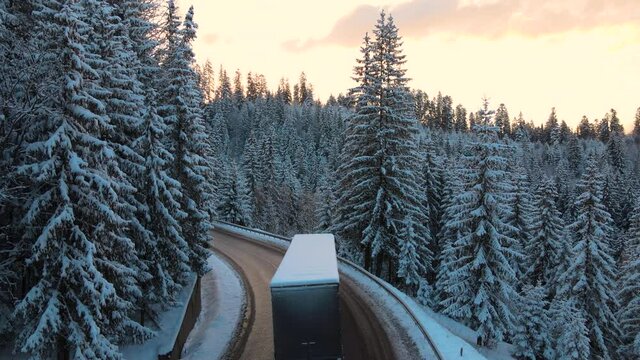 Aerial view of winter landscape with snow covered mountain hills and winding forest road with driving vehicles in morning.