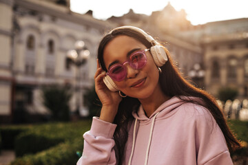Close-up portrait of brunette attractive Asian woman in pink hoodie and sunglasses listening to music in headphones outside.