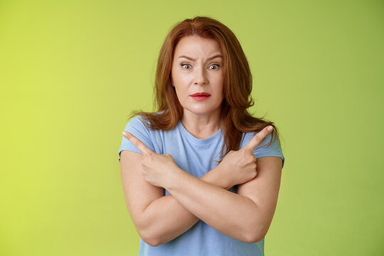 Help Me Choose. Worried Unsure Doubtful Cute Redhead Middle-aged Silly Woman Asking Advice Cross Arms Body Pointing Sideways Left Right Look Concerned Camera Make Difficult Decision Green Background