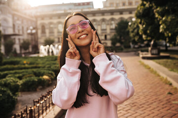 Fototapeta premium Attractive happy tanned girl in pink sunglasses and stylish hoodie smiles, looks up and crosses fingers. Woman poses outside.