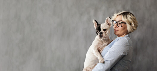 attractive young blonde in glasses with two-tone French bulldog in hands on grey background, model plus size