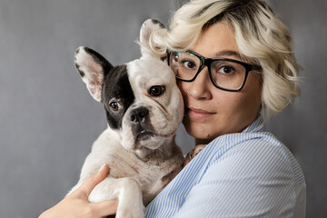 attractive young blonde in glasses with two-tone French bulldog in hands on grey background, model...