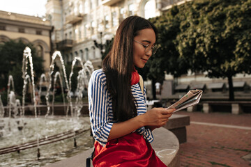 Fototapeta na wymiar Charming tanned woman in striped shirt and red skirt smiles and looks at map. Pretty Asian brunette girl in red headphones poses near fountain.
